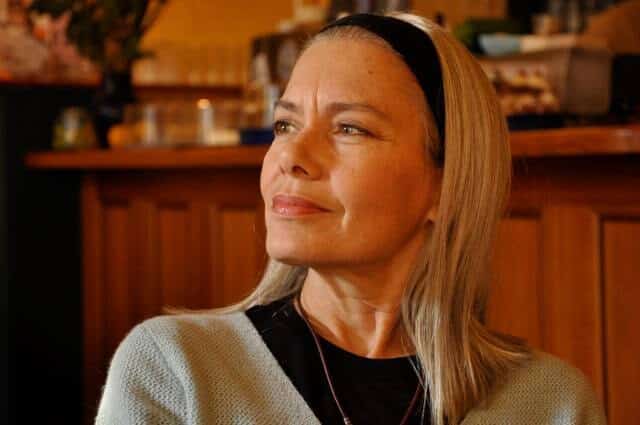 Candida Royalle, cabelos loiros e tiara preta, pensativa e sorridente num café